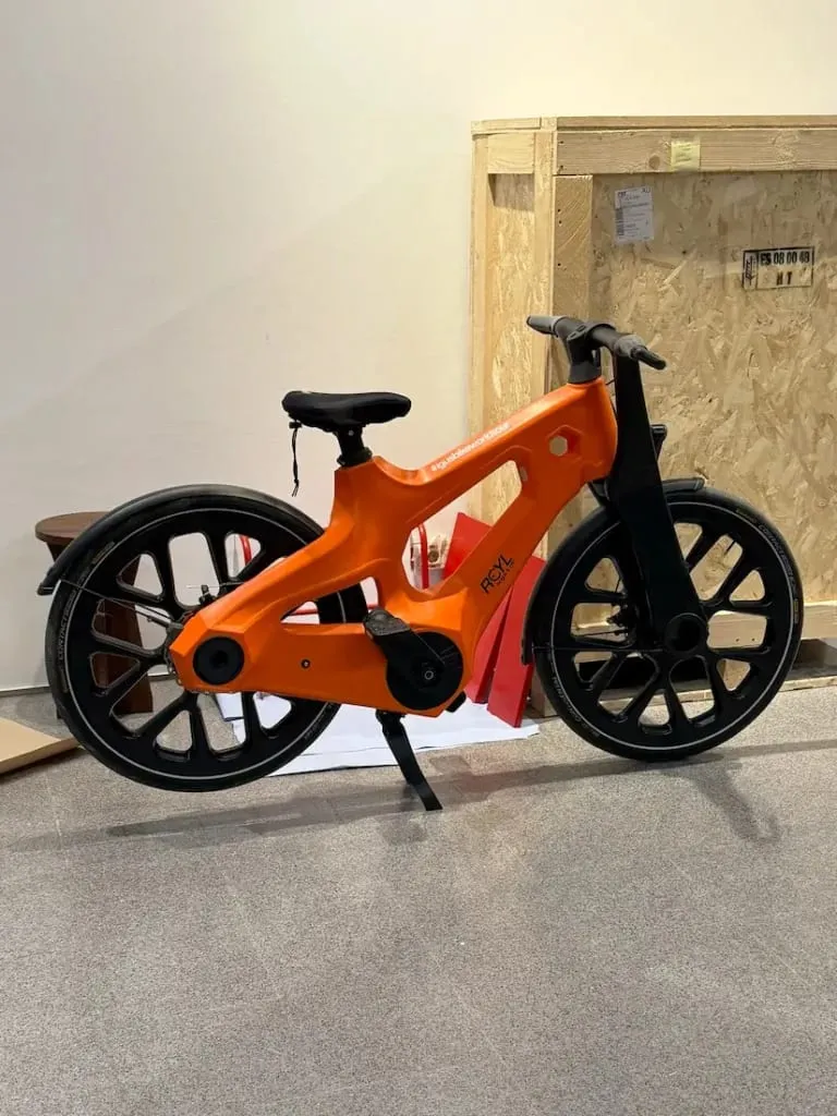 Una bicicleta futurista de color naranja brillante de Igus con ruedas negras gruesas se encuentra en el interior, cerca de una caja de madera, y muestra la innovadora movilidad urbana.