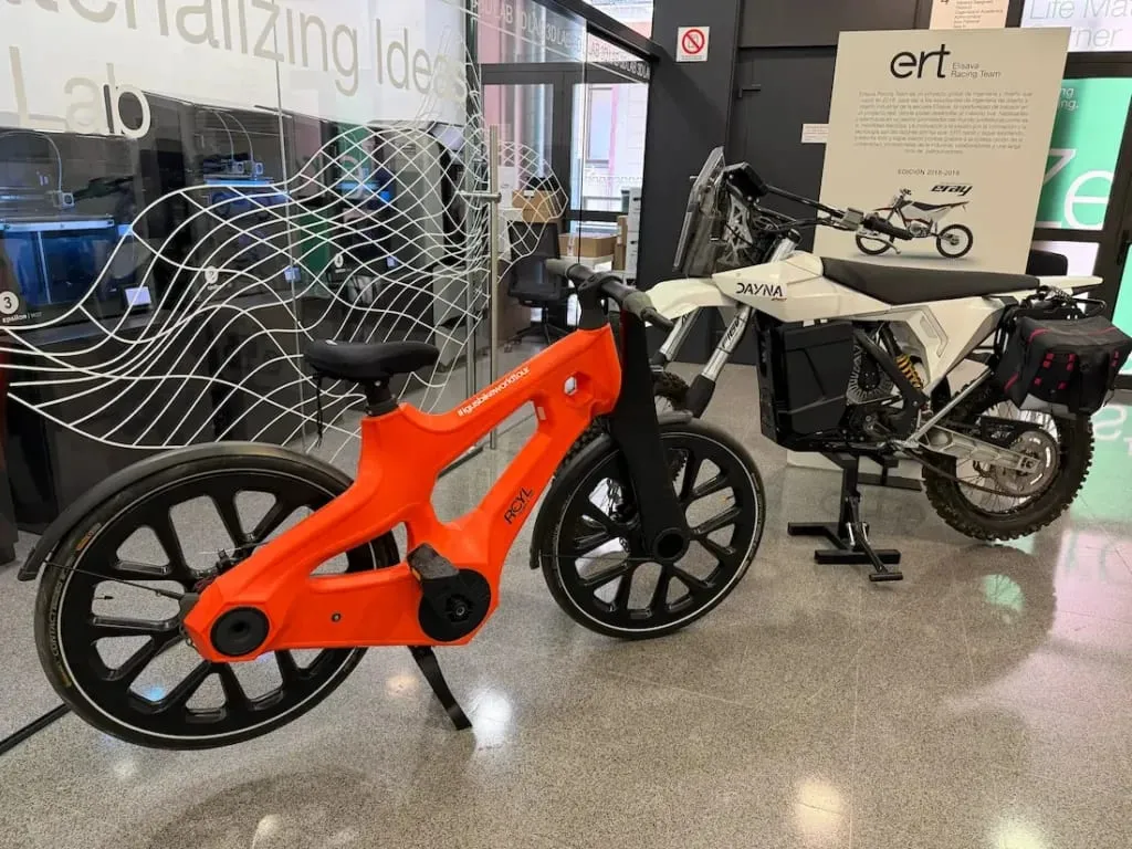 Una bicicleta naranja de igus y una motocicleta eléctrica blanca destacan en la exposición de un moderno espacio, mostrando soluciones innovadoras para la movilidad urbana.