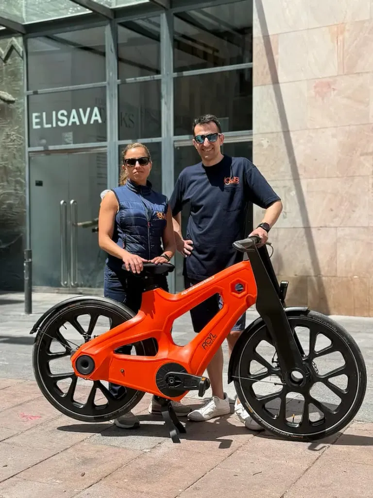 Dos personas se encuentran junto a una bicicleta igus de color naranja brillante en una acera de la ciudad, cerca de la entrada de un edificio, mostrando la innovadora movilidad urbana.