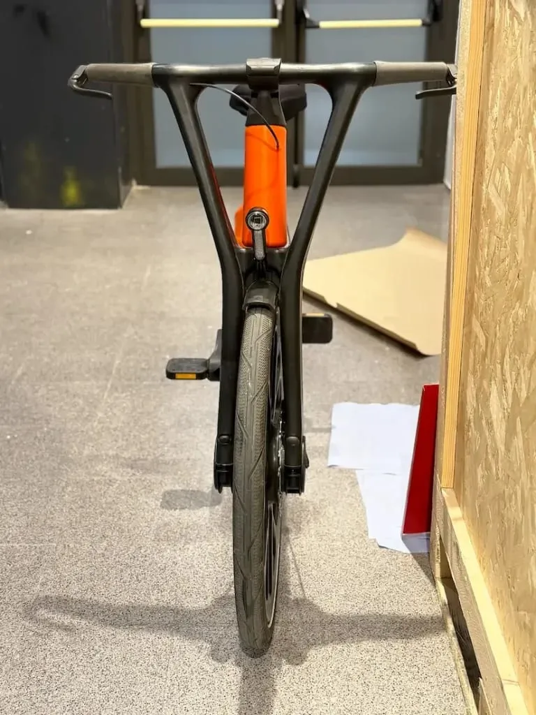 Vista frontal de una bicicleta de igus con cuadro naranja, manillar negro y un único pedal visible, destacando la innovadora movilidad urbana en interiores.