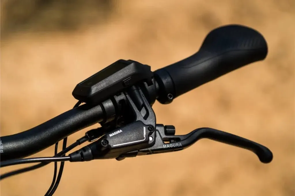 Primer plano de un manillar Riese & Müller Superdelite5 GT rohloff con pantalla digital y palanca de freno Magura, listo para tu próxima aventura, contra un fondo borroso.