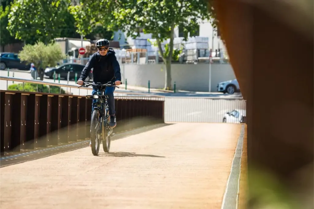 Una persona conduce una Riese & Müller Superdelite5 GT por un puente soleado, enmarcada por árboles y edificios en el fondo, abrazando el espíritu de la aventura.