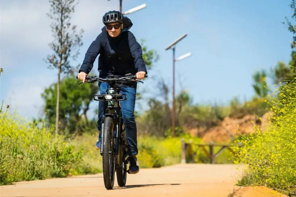 Una persona con casco se embarca en una aventura, conduciendo una bicicleta Riese & Müller Superdelite5 GT rohloff por un camino soleado rodeado de vegetación y flores silvestres amarillas.