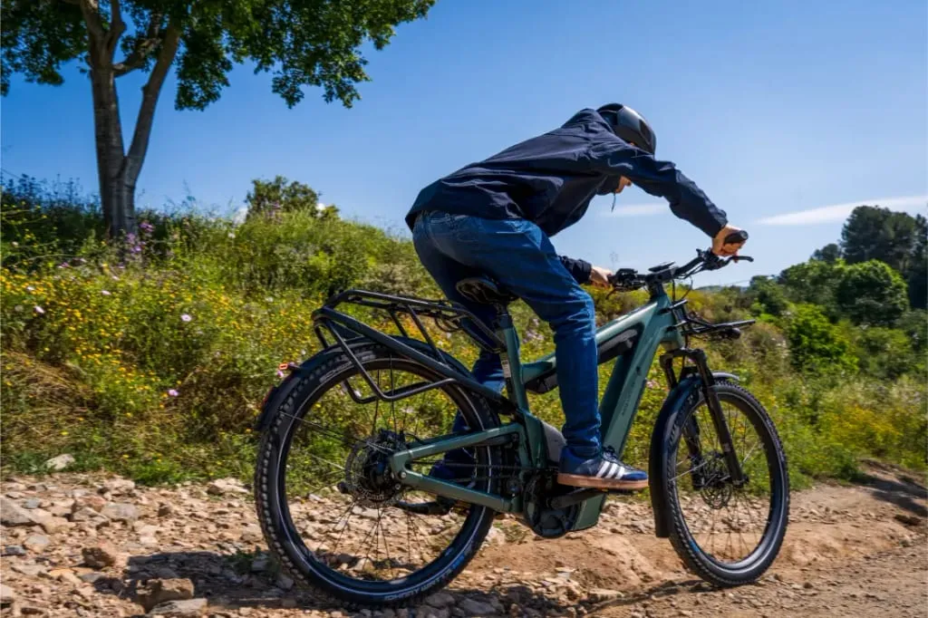 Embárcate en una aventura mientras conduces una bicicleta eléctrica Riese & Müller Superdelite5 GT rohloff cuesta arriba por un camino rocoso, rodeado de exuberante vegetación y árboles bajo un cielo azul claro.