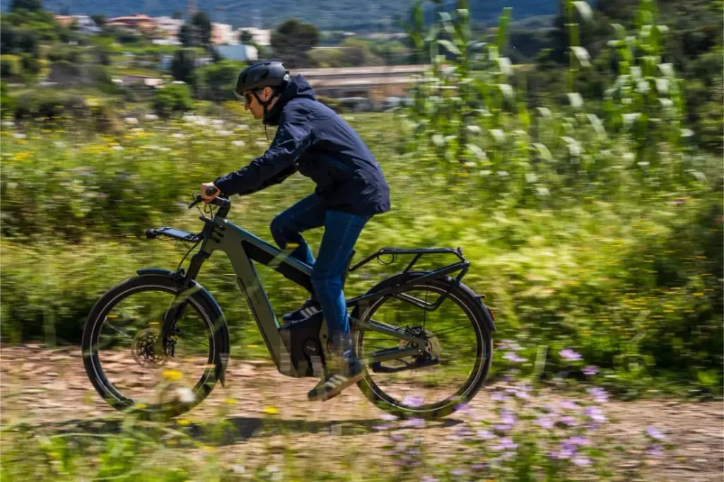 Persona conduciendo una bicicleta eléctrica Riese & Müller Superdelite5 GT rohloff por un camino de tierra rodeado de vegetación y flores silvestres, disfrutando de una aventura por la naturaleza.