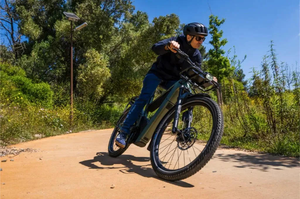 Un hombre con casco y gafas de sol conduce una bicicleta Riese & Müller Superdelite5 GT Rohloff por un camino soleado rodeado de vegetación.
