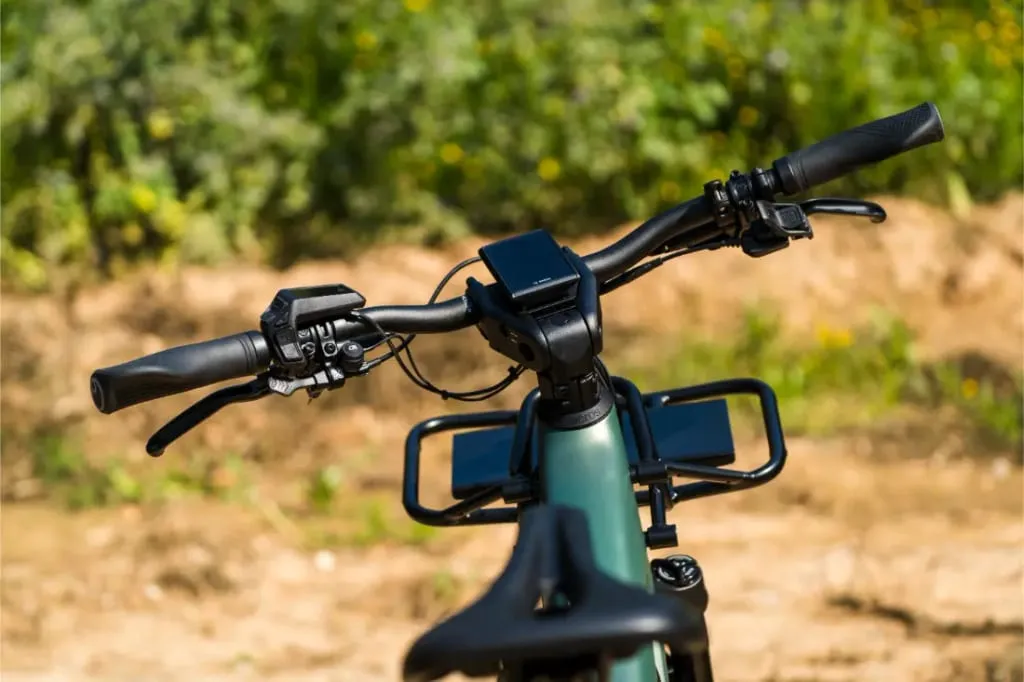 Vista de cerca de un manillar de bicicleta Riese & Müller Superdelite5 GT rohloff con pantallas montadas, al aire libre en una aventura, vegetación y un camino de tierra al fondo.