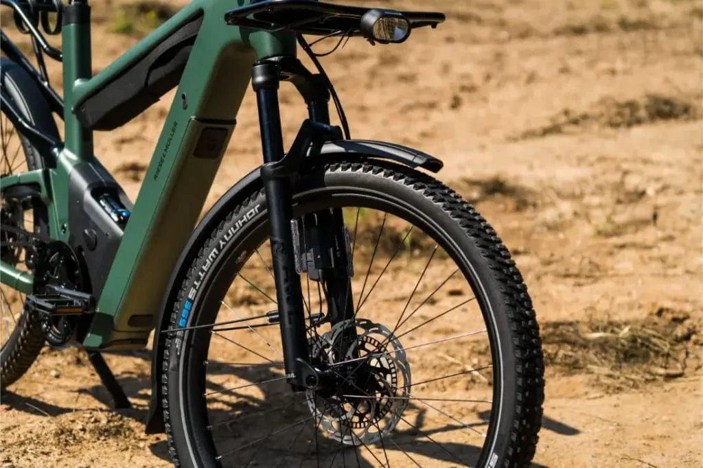 Primer plano de la rueda delantera y la horquilla de una bicicleta eléctrica Riese & Müller Superdelite5 GT rohloff verde sobre un terreno de tierra al aire libre.