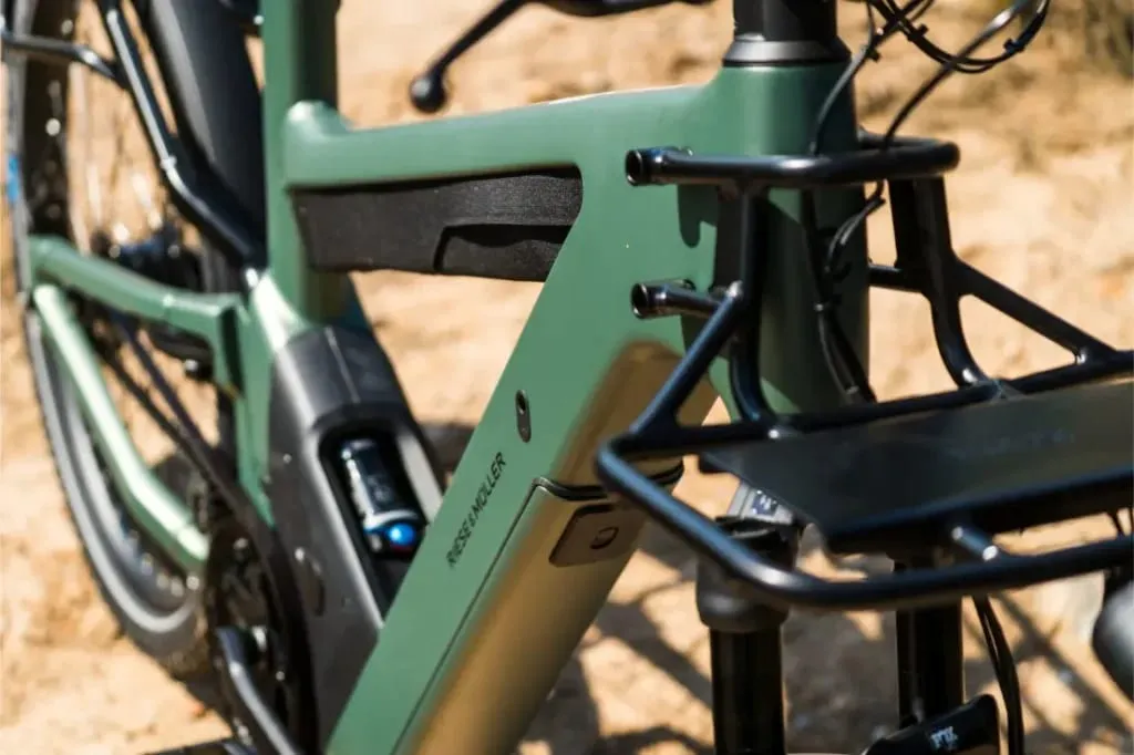 Primer plano del cuadro de una bicicleta eléctrica Riese & Müller Superdelite5 GT verde, que resalta su paquete de batería, sistema Rohloff y portaequipaje frontal al aire libre, perfecto para tu próxima aventura.