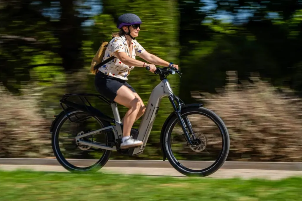 Con casco y mochila, una persona conduce rápidamente una bicicleta eléctrica Riese & Müller Homage5 por un sendero, con vegetación de fondo.