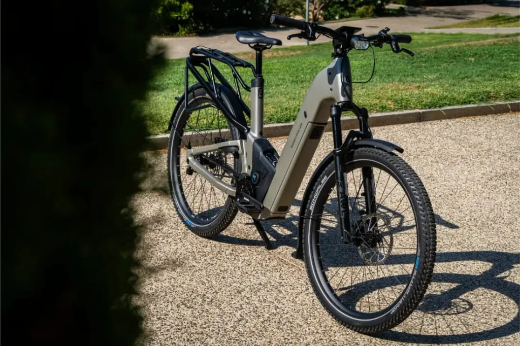 Una moderna bicicleta eléctrica Riese & Müller Homage5 GT con cuadro de paso bajo está aparcada en un camino de grava, rodeada de hierba y árboles al fondo.
