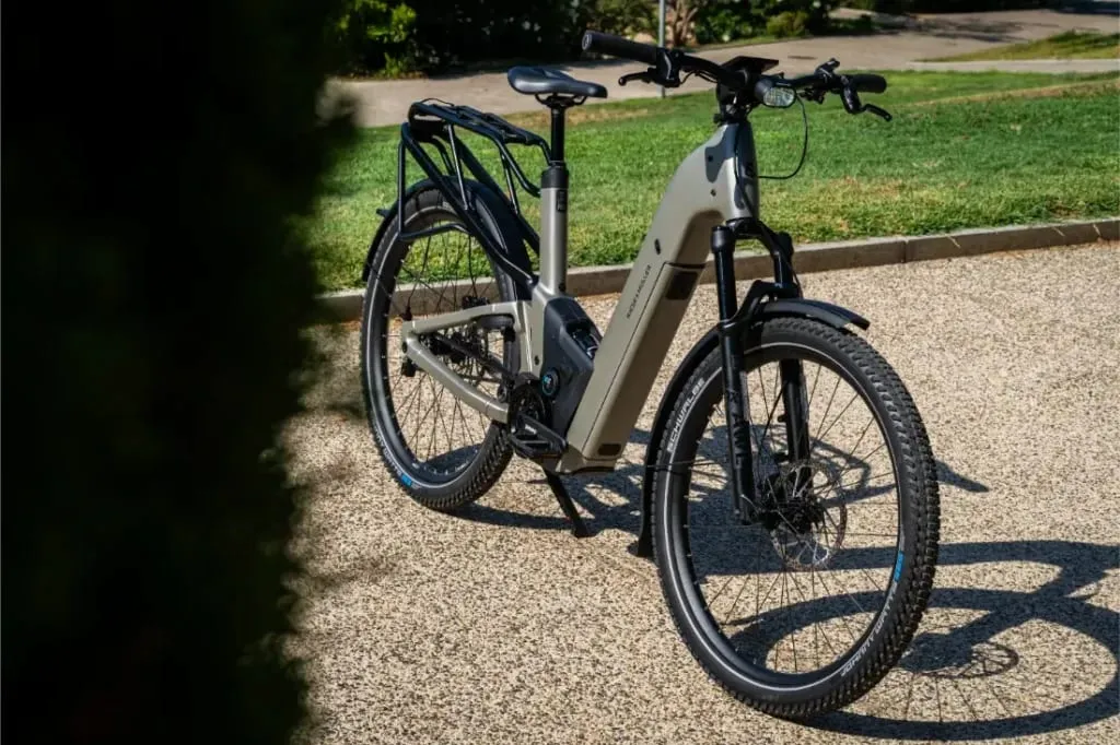 Una moderna bicicleta eléctrica de piñón Riese & Müller Homage5 GT con cuadro de paso bajo está aparcada en un camino de grava, rodeada de hierba y árboles al fondo.