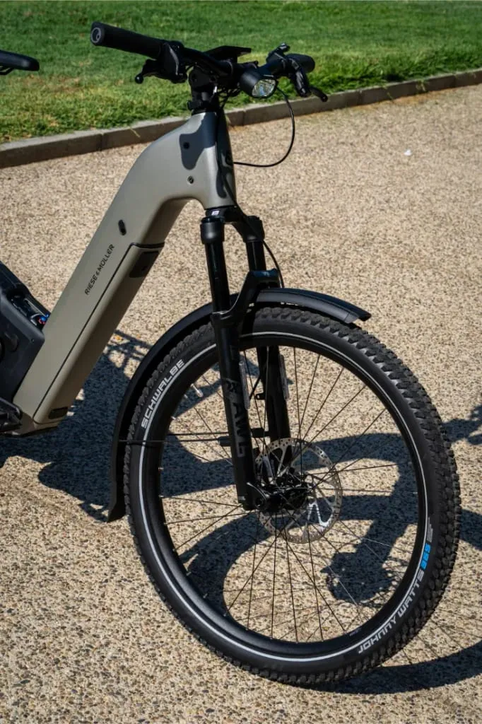 Primer plano de la rueda delantera y el manillar de una bicicleta eléctrica Riese & Müller Homage5 GT Pinion con cuadro de paso bajo, aparcada en un camino de grava.