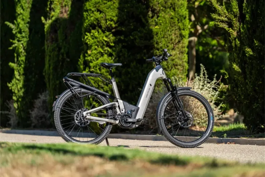 Una moderna bicicleta eléctrica Riese & Müller Homage5 con portabultos trasero, con diseño de paso bajo, aparcada en un camino pavimentado junto a árboles y arbustos verdes.