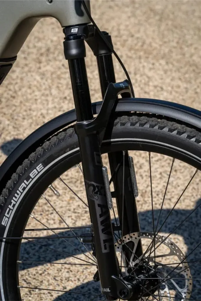 Primer plano de una horquilla de suspensión delantera de bicicleta, neumático Schwalbe y freno de disco sobre una superficie de grava, similar a la robusta configuración que se encuentra en la Riese & Müller Homage5 paso bajo.