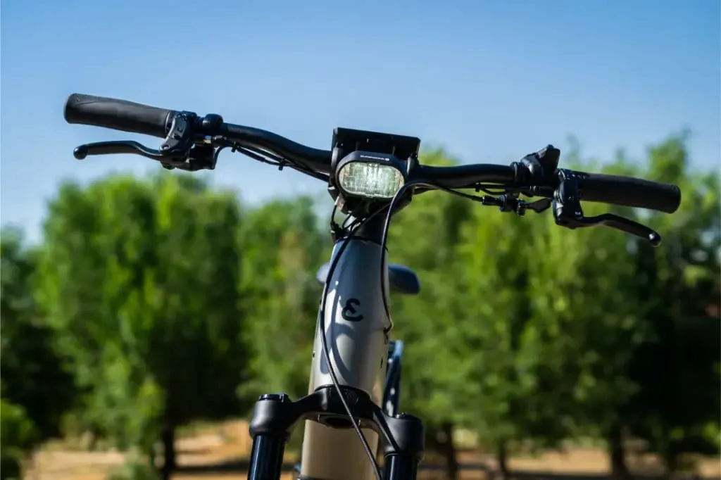 Primer plano del manillar y el faro de una bicicleta eléctrica Riese & Müller Homage5, con árboles y cielo azul de fondo.