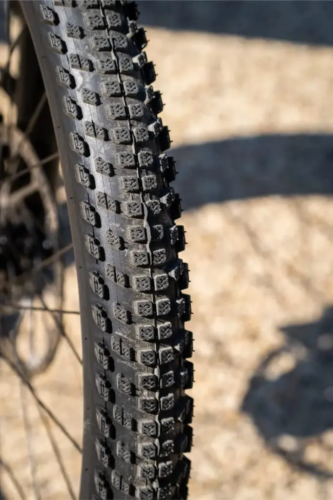 Primer plano de un neumático GT de piñón de bicicleta de montaña con dibujo de tacos, iluminado por la luz del sol sobre una superficie de grava.