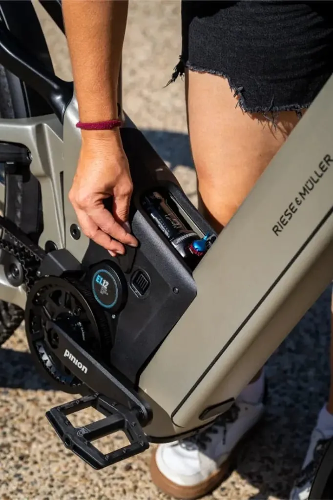 Una persona en pantalón corto y zapatillas blancas coloca o quita una batería de una bicicleta eléctrica Riese & Müller Homage5 GT con cuadro de paso bajo.