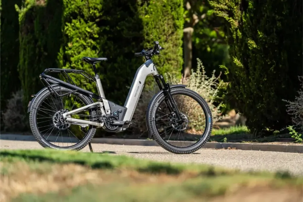 Una bicicleta eléctrica blanca Riese & Müller Homage5 está aparcada en un camino pavimentado con árboles verdes al fondo.