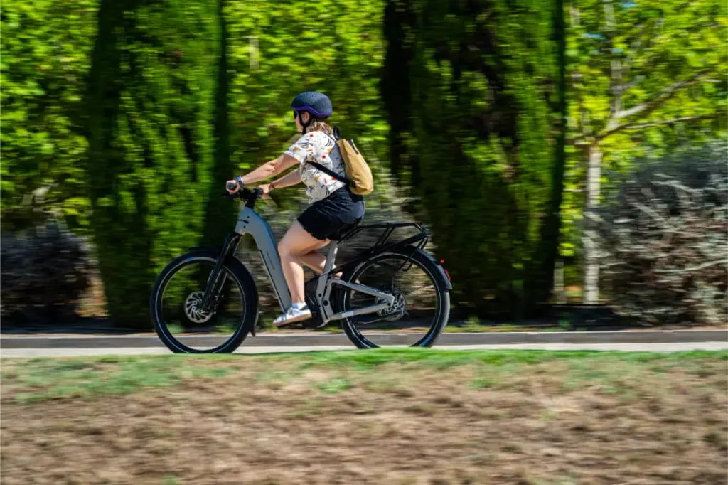 Con casco, una persona monta en una Riese & Müller Homage5 con paso bajo por un sendero bordeado de árboles bajo el sol.