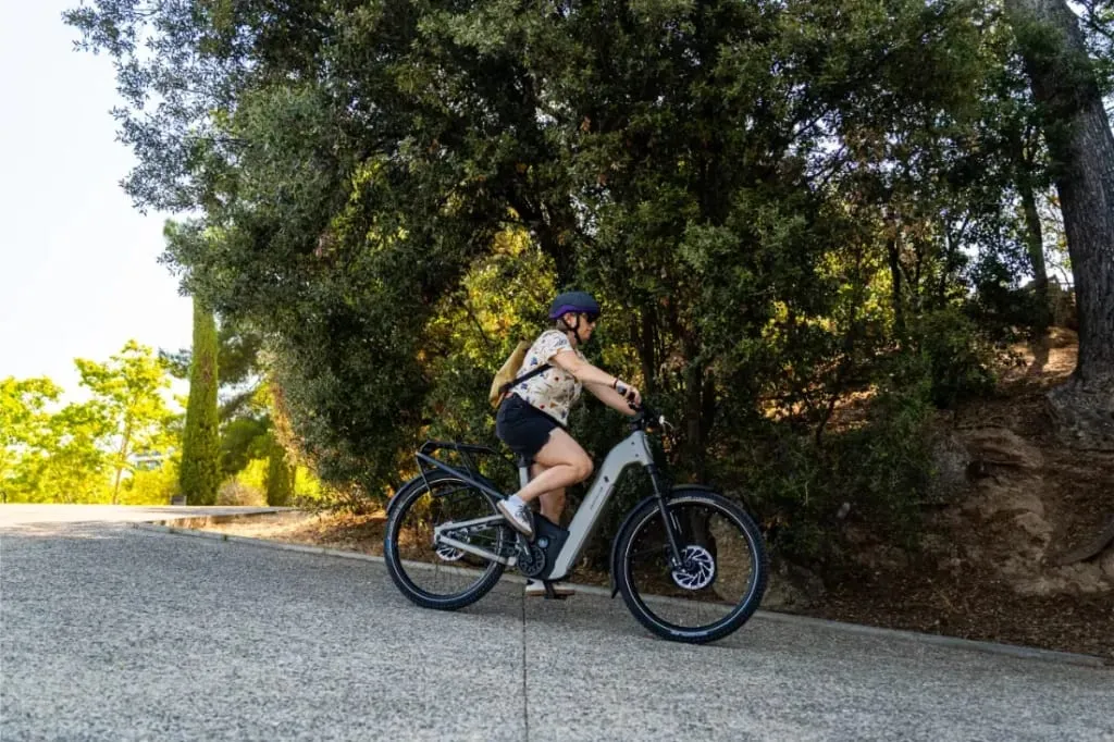 Una persona monta en una bicicleta eléctrica de piñón Riese & Müller Homage5 GT con paso bajo cuesta arriba por un camino pavimentado, rodeado de frondosos árboles y vegetación.
