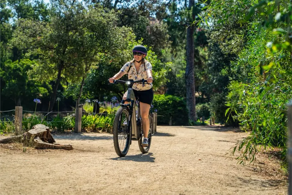 Una persona con casco monta en una bicicleta Riese & Müller Homage5 GT Pinion con paso bajo por un soleado camino de tierra rodeado de árboles y plantas verdes.
