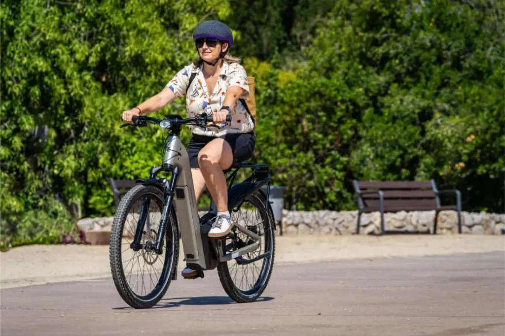 Mujer con casco y gafas de sol monta una e-bike Riese & Müller Homage5 GT con paso bajo y piñón en un día soleado en el parque.