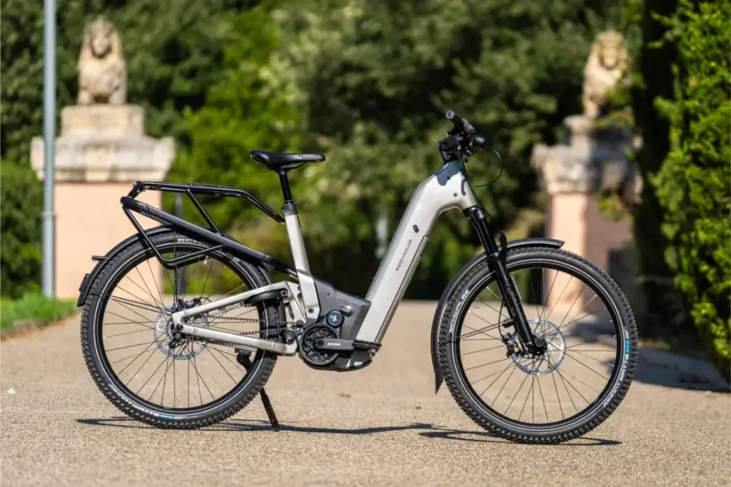 Una bicicleta eléctrica Riese & Müller Homage5 plateada con cuadro de paso bajo está aparcada en un camino, rodeada de vegetación y estatuas de leones de piedra al fondo.