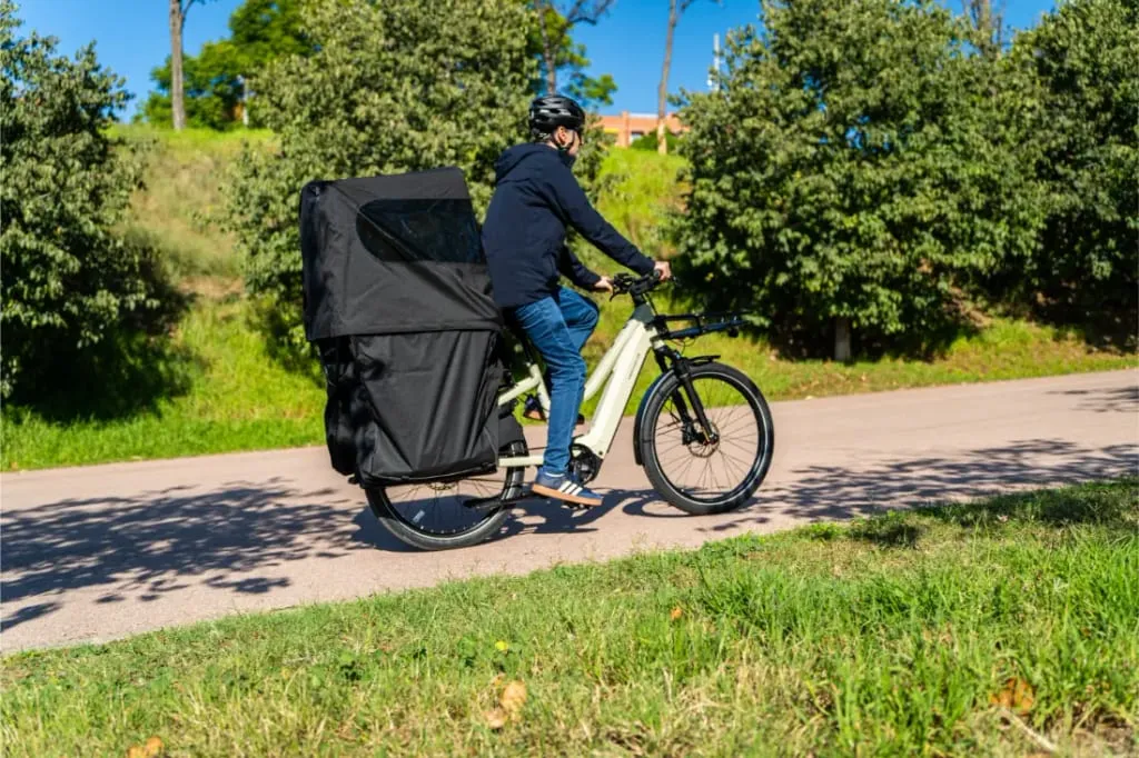 Una persona monta en una bicicleta de carga con una gran cubierta de carga negra acoplada, pedaleando por una soleada carretera bordeada de vegetación.