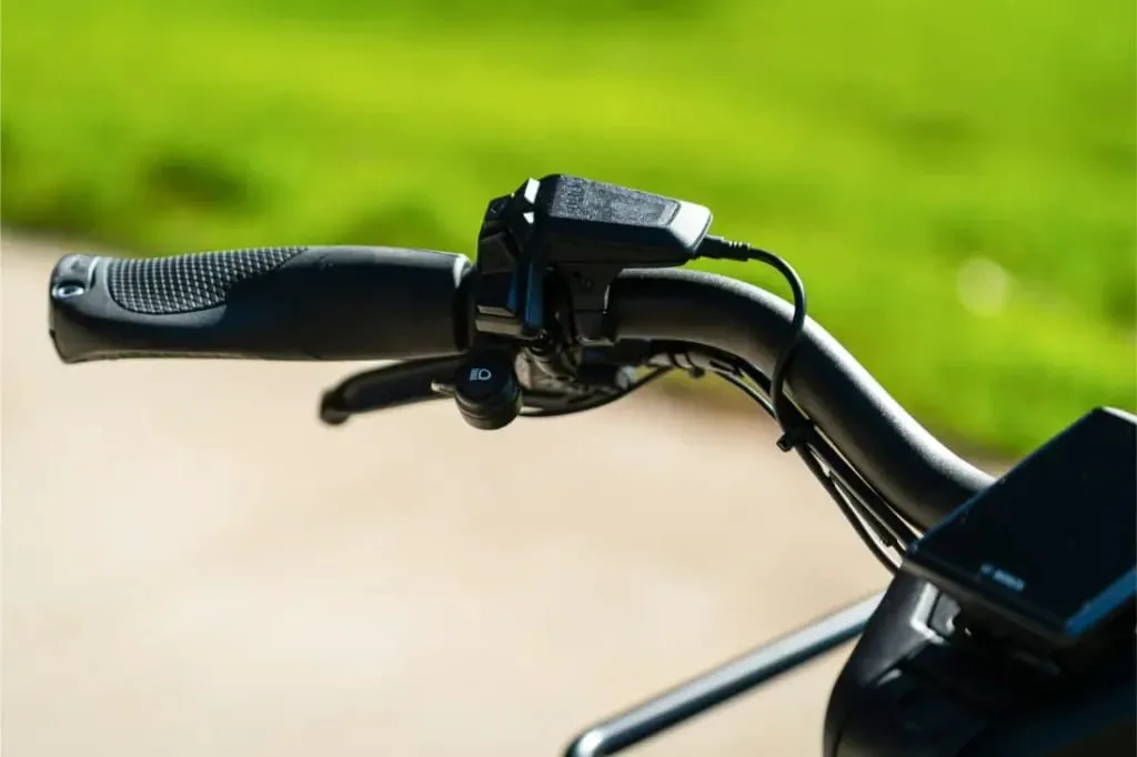 Primer plano de un manillar Riese & Müller Multicharger3 Mixte Vario con un dispositivo electrónico, con hierba verde suavemente difuminada en el fondo, destacando sus prácticas capacidades de carga.