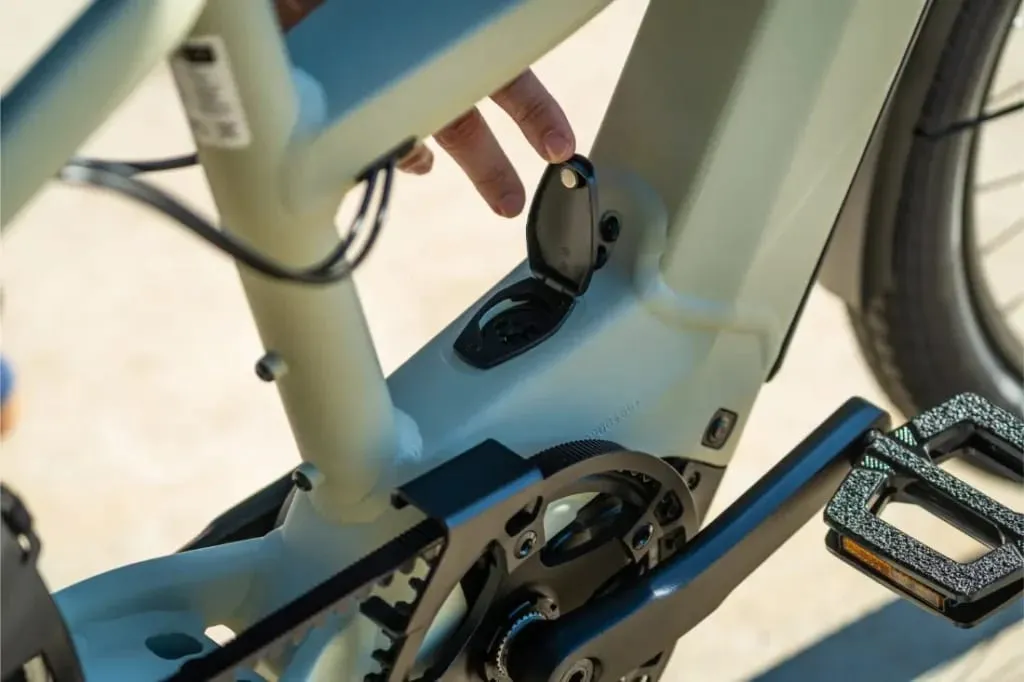 Una mano abre la tapa del compartimento de la batería en un cuadro Riese & Müller Multicharger3 Mixte Vario cerca del pedal, preparándose para la carga.