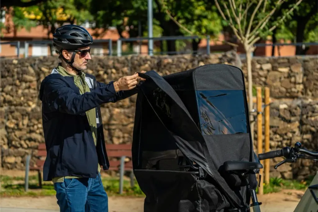 Un hombre con casco ajusta la cubierta de un remolque de bicicleta negro Riese & Müller Multicharger3 Mixte Vario, preparándose para una carga seria, al aire libre en un día soleado.