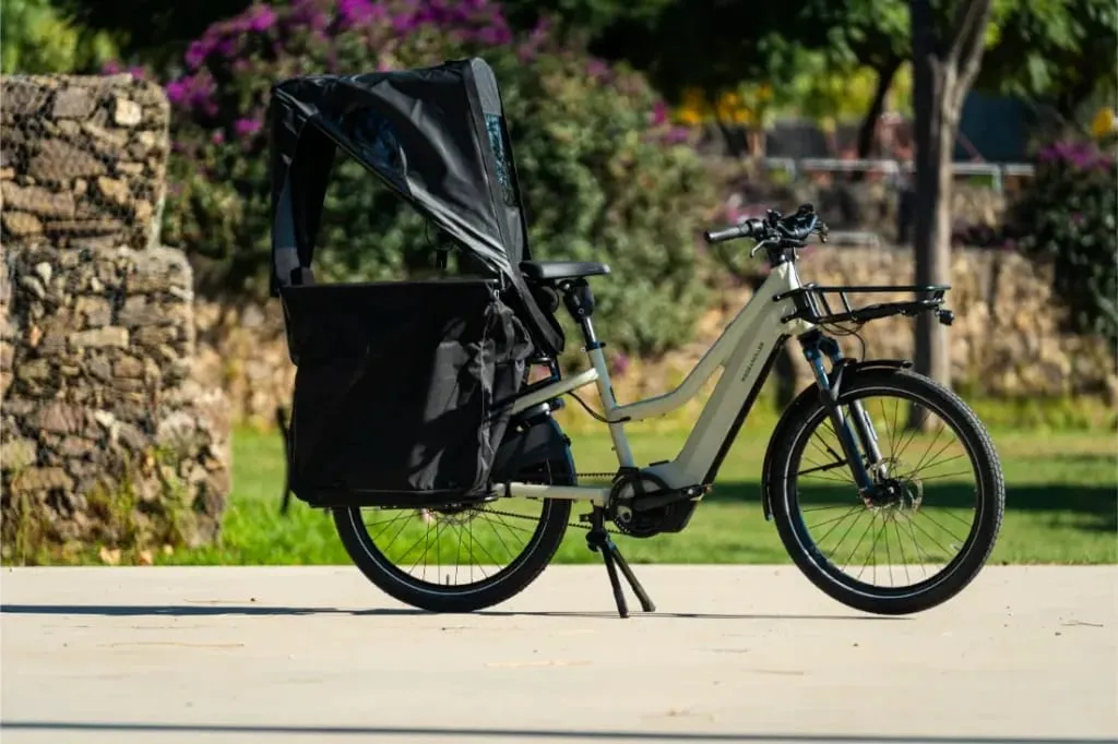 Una bicicleta eléctrica de carga Riese & Müller Multicharger3 con una gran capota negra y una cesta está aparcada al aire libre en un día soleado.