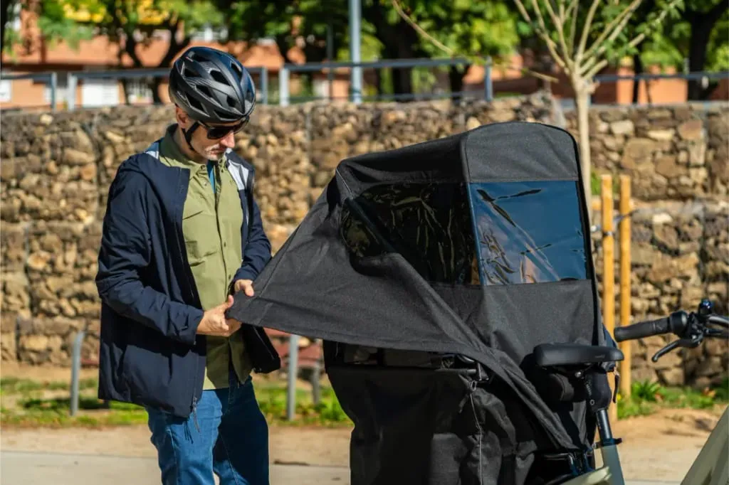 Con casco, una persona ajusta la cubierta de un asiento infantil de una bicicleta de carga Riese & Müller Multicharger3 Mixte Vario al aire libre en un día soleado.