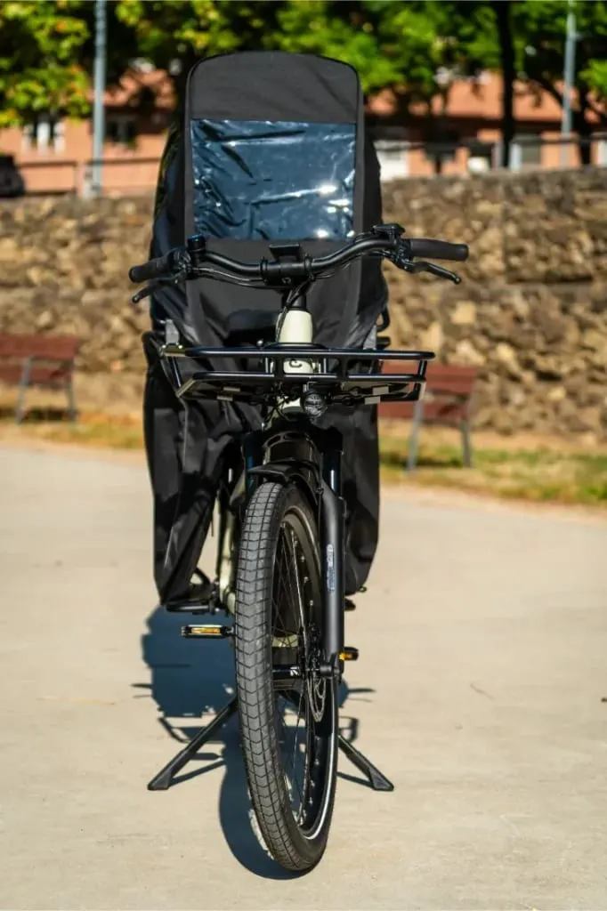 Vista frontal de una bicicleta de carga Riese & Müller Multicharger3 Mixte Vario con asiento para niños, protegida por una cubierta de plástico negro y transparente para la lluvia.