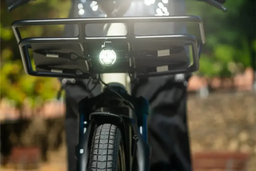 Primer plano de una rueda delantera, faro delantero y portacargas Riese & Müller Multicharger3 a la luz del sol, mostrando esta versátil bicicleta de carga.