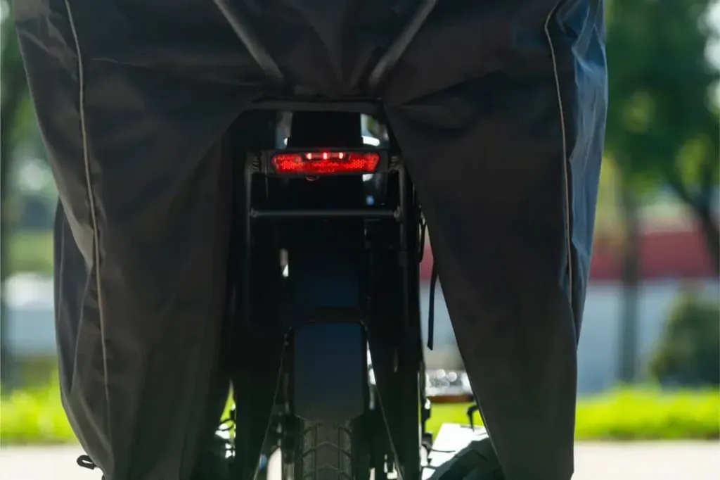 Vista trasera de una bicicleta eléctrica cubierta Riese & Müller Multicharger3 Mixte Vario con su piloto trasero rojo iluminado, al aire libre a la luz del día, lista para su próxima carga.