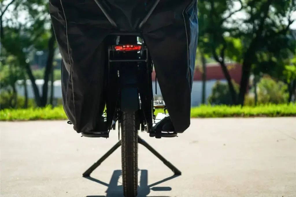 Vista trasera de una bicicleta eléctrica Riese & Müller Multicharger3 cubierta, con la pata de cabra bajada, aparcada en la acera y enmarcada por una exuberante vegetación al fondo.