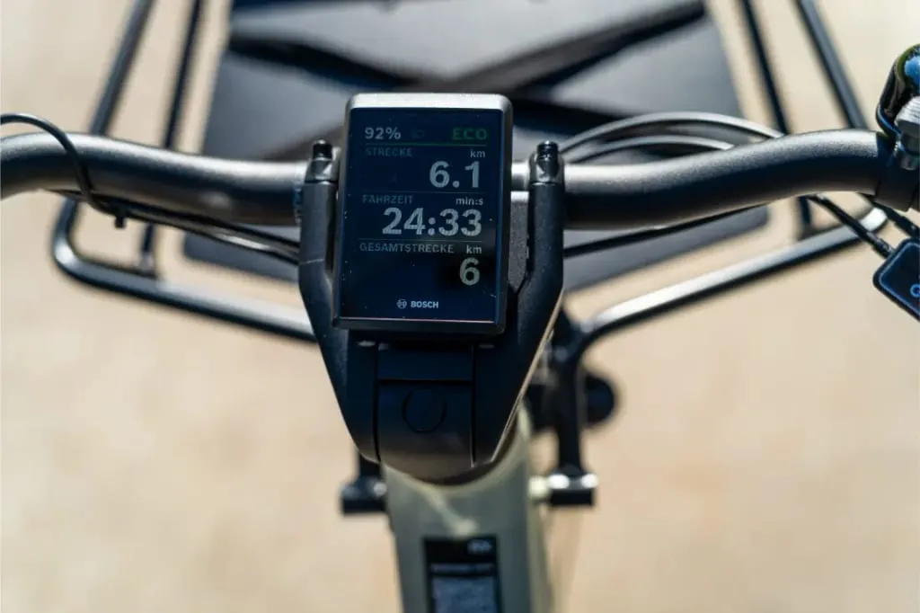 Primer plano de un manillar Riese & Müller Multicharger3 Mixte Vario con una pantalla digital que muestra la velocidad, la distancia y el estado de la batería, destacando sus características avanzadas para la carga diaria y la comodidad.