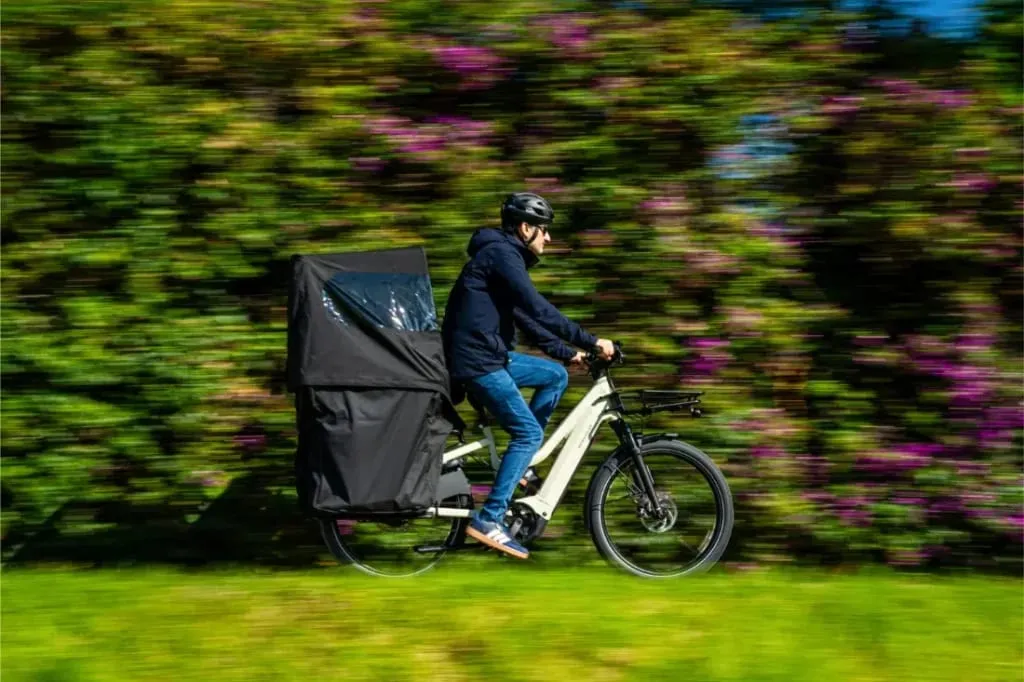 Persona con casco conduce una bicicleta de carga Riese & Müller Multicharger3 Mixte Vario con una gran caja negra, pasando junto a vegetación y flores moradas.