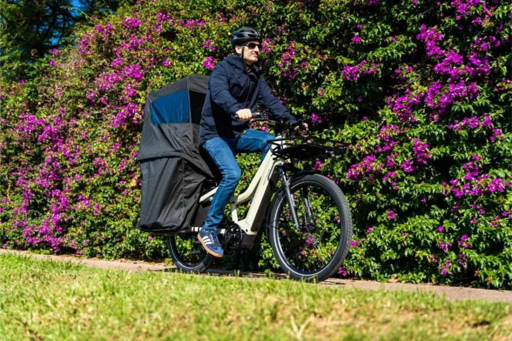 Un hombre monta un Riese & Müller Multicharger3 Mixte Vario con un asiento infantil cubierto, pasando junto a un seto de flores púrpuras en un día soleado, una escena perfecta para aventuras de carga familiar.