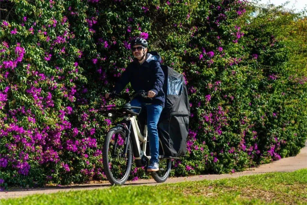 Una persona montada en una Riese & Müller Multicharger3 Mixte Vario con una gran mochila negra de carga, pasa junto a una pared cubierta de vibrantes flores moradas.