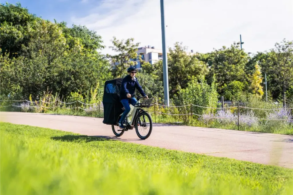 Una persona monta en una Riese & Müller Multicharger3 Mixte Vario, una versátil bicicleta de carga con una gran bolsa de carga, a lo largo de un soleado y verde sendero del parque.