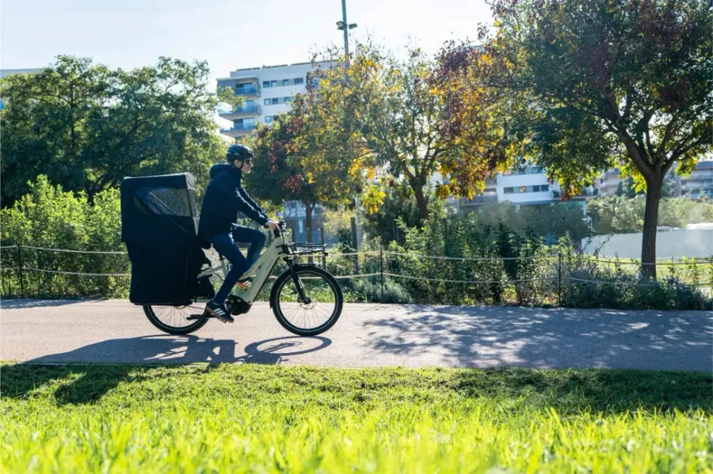 Una persona conduce un Riese & Müller Multicharger3 Mixte Vario con asiento cubierto para el pasajero por un soleado sendero de un parque, rodeado de árboles y hierba, lo que lo hace perfecto para transportar carga en aventuras al aire libre.