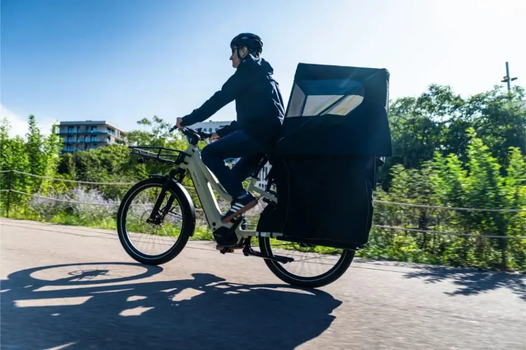 Una persona monta en una bicicleta de carga Riese & Müller Multicharger3 con un gran compartimento cubierto en un día soleado, árboles y edificios al fondo.