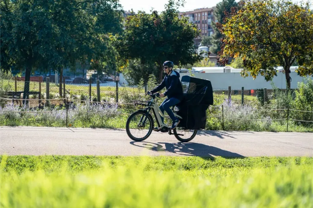 Persona montando en una e-bike Riese & Müller Multicharger3 Mixte Vario con una bolsa de carga grande por el sendero de un parque, rodeado de vegetación.