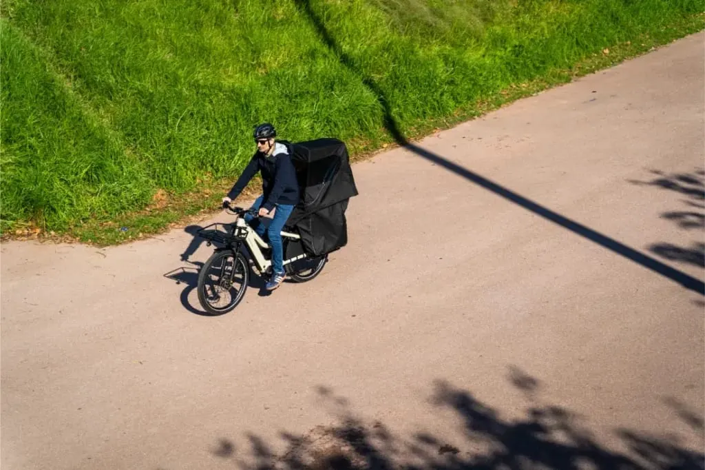 Una persona conduce una bicicleta eléctrica de carga Riese & Müller Multicharger3 Mixte Vario con una caja de entrega negra por una carretera soleada junto a hierba verde.