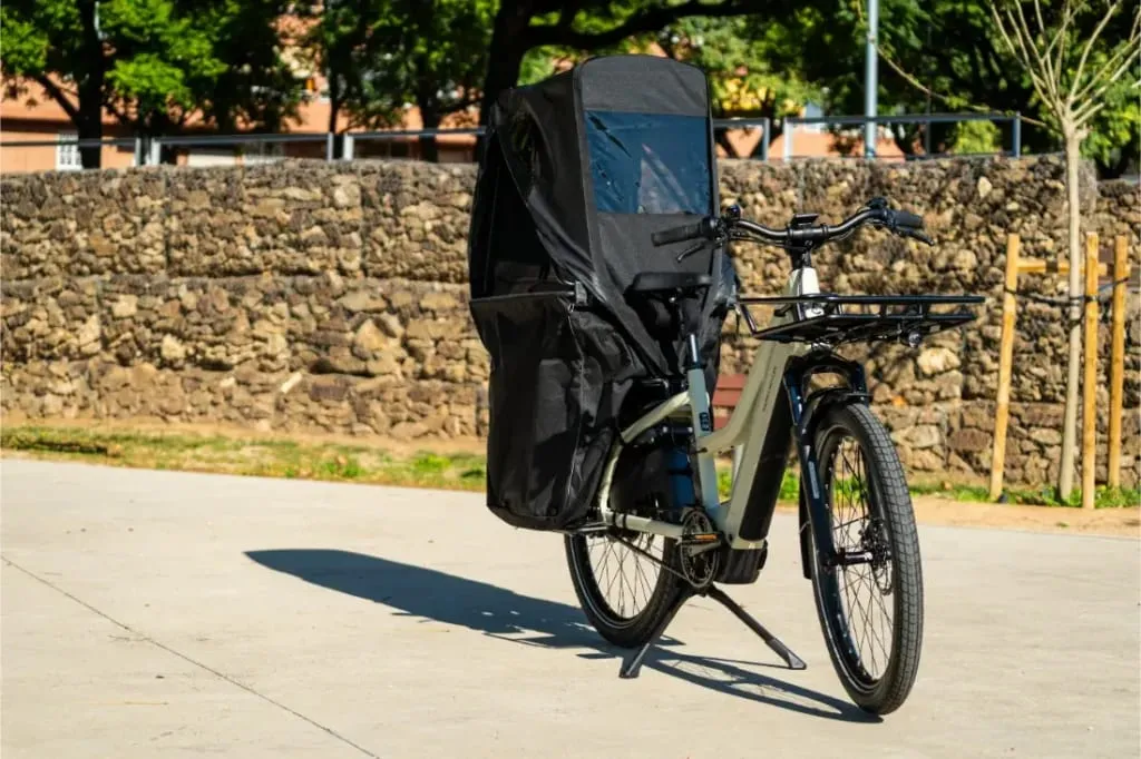 Una bicicleta negra Riese & Müller Multicharger3 Mixte Vario con un gran asiento infantil y cubierta para la lluvia está aparcada en una soleada zona pavimentada cerca de un muro de piedra, lista para cualquier carga o aventura familiar.