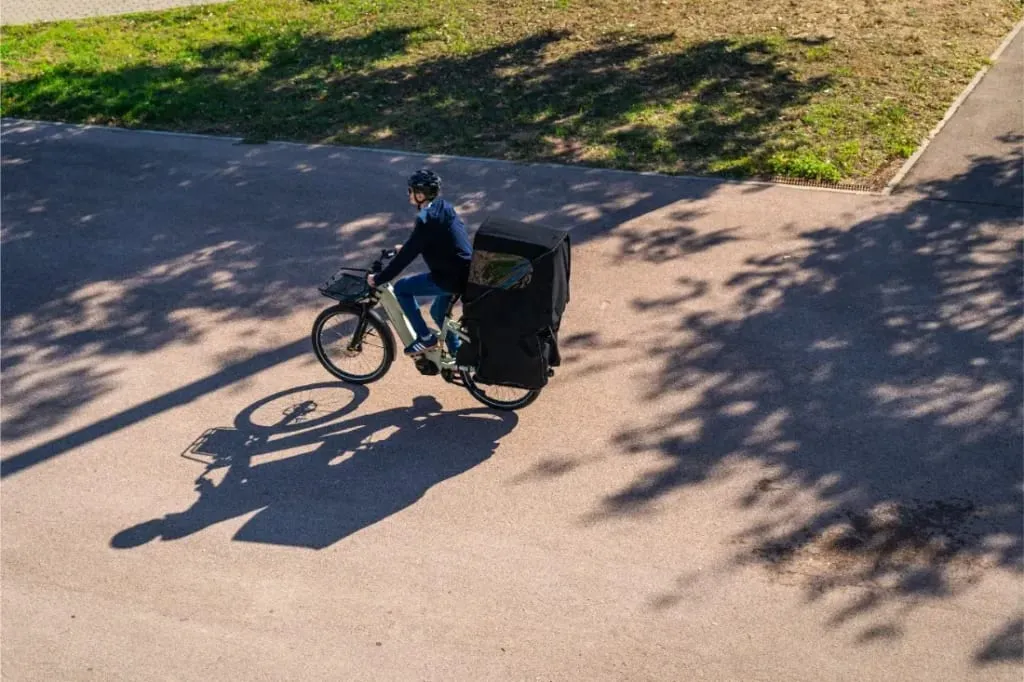Una persona monta en una bicicleta Riese & Müller Multicharger3 Mixte Vario por una carretera iluminada por el sol, llevando a la espalda una gran bolsa de carga negra.