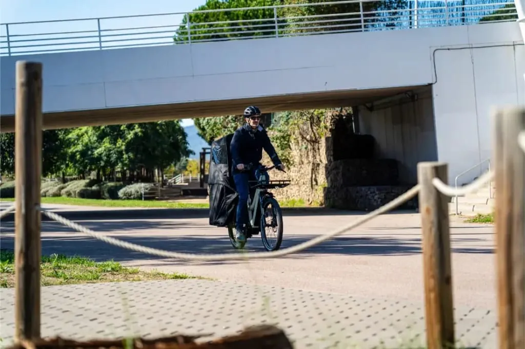 Persona con casco monta una bicicleta de carga Riese & Müller Multicharger3 Mixte Vario con una gran bolsa negra debajo de un puente en un día soleado.